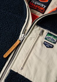 Giacca in pile con zip caratterizzata da sezioni blu navy e crema, superficie testurizzata, doppia zip e toppe con logo in colori a contrasto.