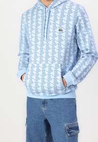 Osoba v světle modré mikině s geometrickým vzorem a přední kapsou, v modrých džínách se postranní kapsou, s rukama v kapsách mikiny.