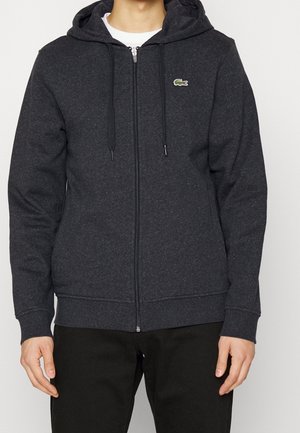 Homme portant un sweat à capuche zippé gris foncé avec un petit logo crocodile vert sur la poitrine, associé à un pantalon noir.