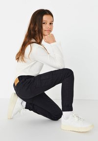 Calças de trabalho pretas com punhos elásticos, bolsos laterais e uma etiqueta de couro na parte de trás. Combinadas com ténis brancos e uma blusa branca de manga longa.