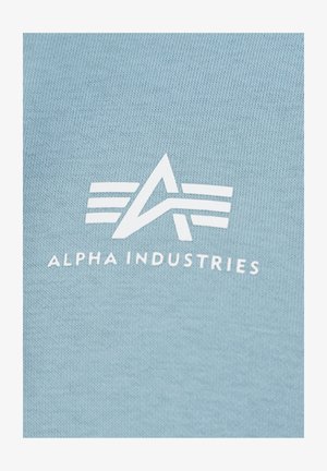 Svijetloplavi pamučni materijal s istaknutim bijelim logotipom geometrijskog dizajna i natpisom "ALPHA INDUSTRIES" ispod logotipa.