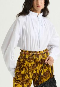 Camicia bianca a maniche lunghe con collo alto; abbinata a pantaloni gialli con motivo floreale e cintura decorativa con accenti colorati.