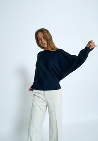 Marineblauer, gestrickter Pullover mit weitem Bootsausschnitt und fällt Schulter, kombiniert mit hellbeigen, weit geschnittenen Hosen. Glatte, weiche Textur.