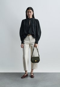 Γυναίκα που στέκεται φορώντας μαύρο μπλουζάκι με μακριά μανίκια, μπεζ παντελόνι, μπορντό φλατ παπούτσια, κρατώντας μια μικρή σκούρα πράσινη τσάντα στο πλευρό της.