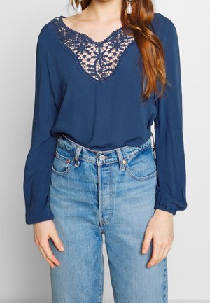 Blusa a maniche lunghe blu navy con colletto in pizzo all'uncinetto, abbinata a jeans in denim blu chiaro a vita alta. La mano del modello poggia sui fianchi.