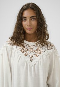 Blusa bianca con dettagli in pizzo intricati attorno al collo, maniche ampie e una texture di tessuto morbido e leggero.