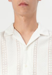 Camisa blanca de manga corta con un patrón texturizado, cierre de botones y un collar clásico con acentos bordados a lo largo de líneas verticales.