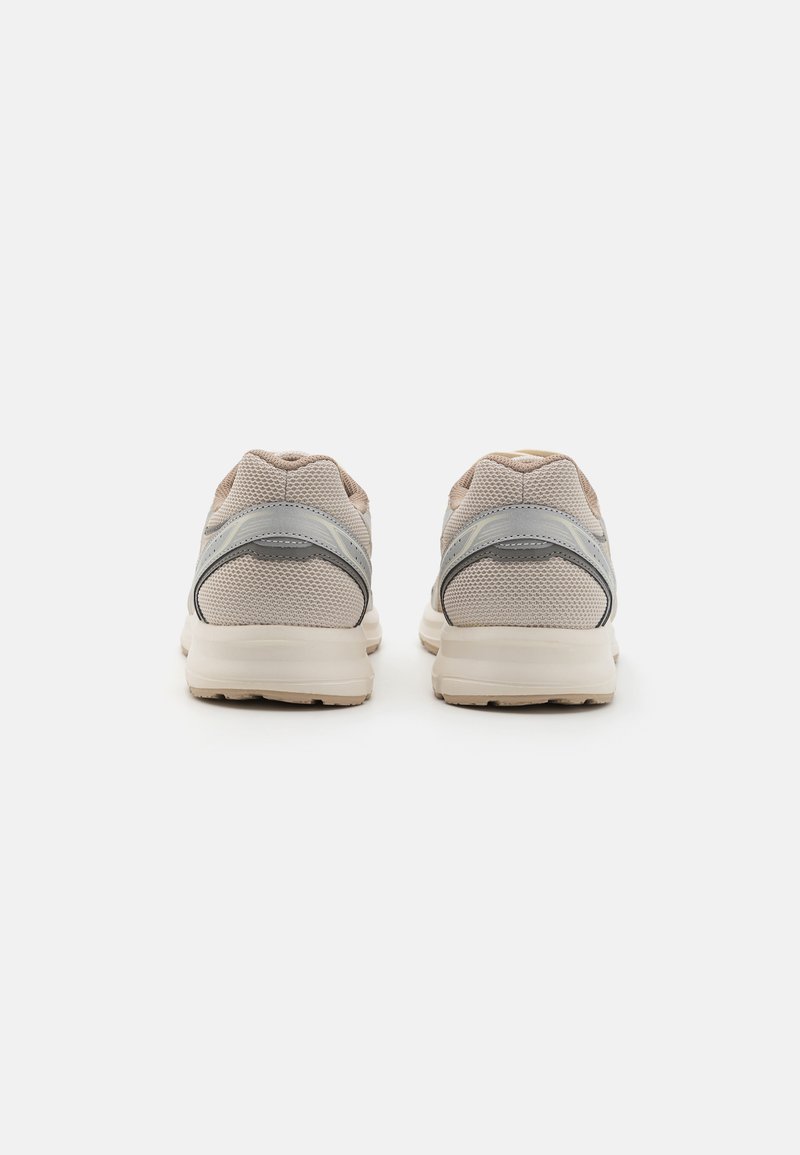 Zapatillas deportivas de malla beige con un acabado texturizado, que presentan detalles en gris y un collar acolchado. La suela es de tono claro y ligeramente curvada.