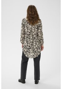Chemisier à manches longues avec un motif de feuilles noir et crème, en tissu transparent, à ourlet arrondi et coupe décontractée, associé à un pantalon noir et des chaussures.