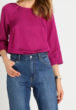 Mujer que lleva una blusa de manga larga magenta metida en unos vaqueros azules de talle alto con un reloj de pulsera plateado en el brazo derecho.