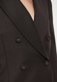 Blazer de doble botonadura en mezcla de lana marrón oscuro, con grandes botones redondos y cuello con muesca. Textura suave con un ajuste entallado.