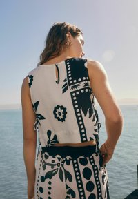 Witte cropped top met zwarte bloemen- en geometrische patronen, heeft een kleine sleutelgaatje-opening aan de achterkant en strikjes aan de zijkanten.