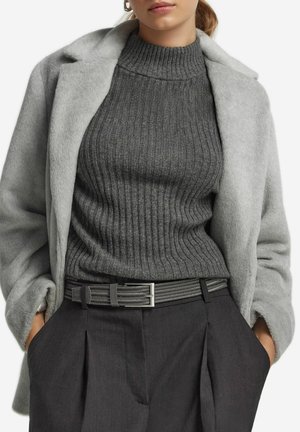 Femme portant un manteau en fausse fourrure gris clair sur un pull col roulé côtelé gris foncé, rentré dans un pantalon plissé gris foncé avec une ceinture grise.