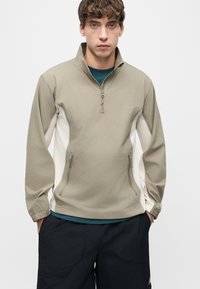 Νεαρός άνδρας που φοράει μπεζ και λευκό μπουφάν με φερμουάρ στο ένα τέταρτο και πλαϊνές τσέπες, σκούρο παντελόνι, που στέκεται μπροστά σε απλό φωτεινό φόντο.