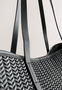 Sac cabas noir avec une texture tissée, des poignées brillantes et un design robuste. Présente un motif géométrique sur la surface principale.