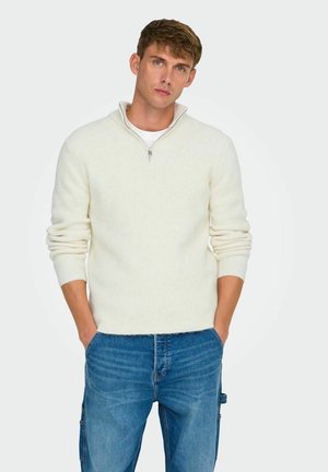 Pullover sweater in soft cream knit with a half-zip design, featuring a ribbed texture and long sleeves paired with blue denim jeans.