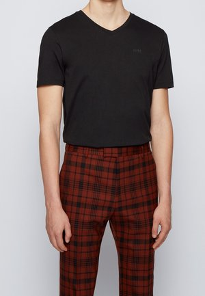 Mand iført sort T-shirt med korte ærmer og V-hals samt højtaljede røde og sorte ternede bukser, stående foran en ensfarvet baggrund.