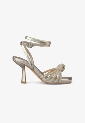 Sandale à talons hauts en métal argenté avec un bout arrondi, une délicate sangle à la cheville et des accents décoratifs en nœuds. Les brides texturées ajoutent du détail.