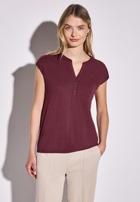 Bordeaux kortærmet top med henley-udskæring og tre knapper. Letvægtsstof med løs pasform, parret med beige bukser.
