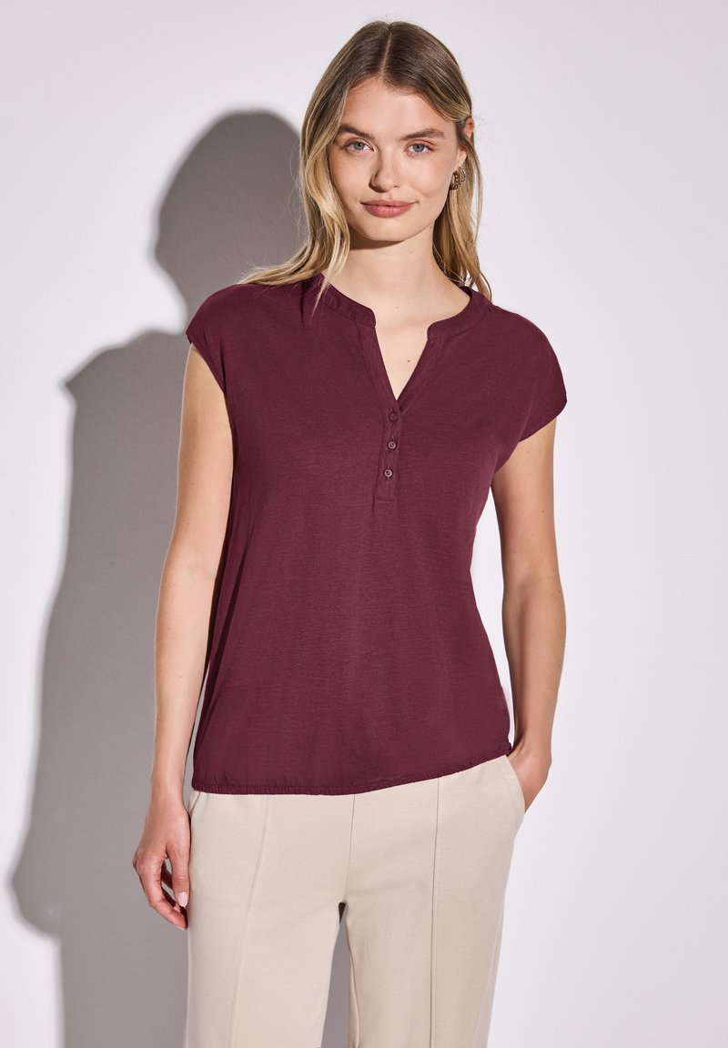 Bordeaux kortærmet top med henley-udskæring og tre knapper. Letvægtsstof med løs pasform, parret med beige bukser.