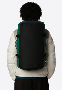 Sac à dos noir et vert de forme rectangulaire, en tissu, avec des bretelles ajustables et des boucles latérales, porté sur une veste blanche en duvet.