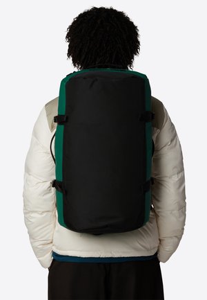 Zaino nero e verde di forma rettangolare, in materiale tessile, con spallacci regolabili e fibbie laterali, indossato sopra una giacca bianca imbottita.