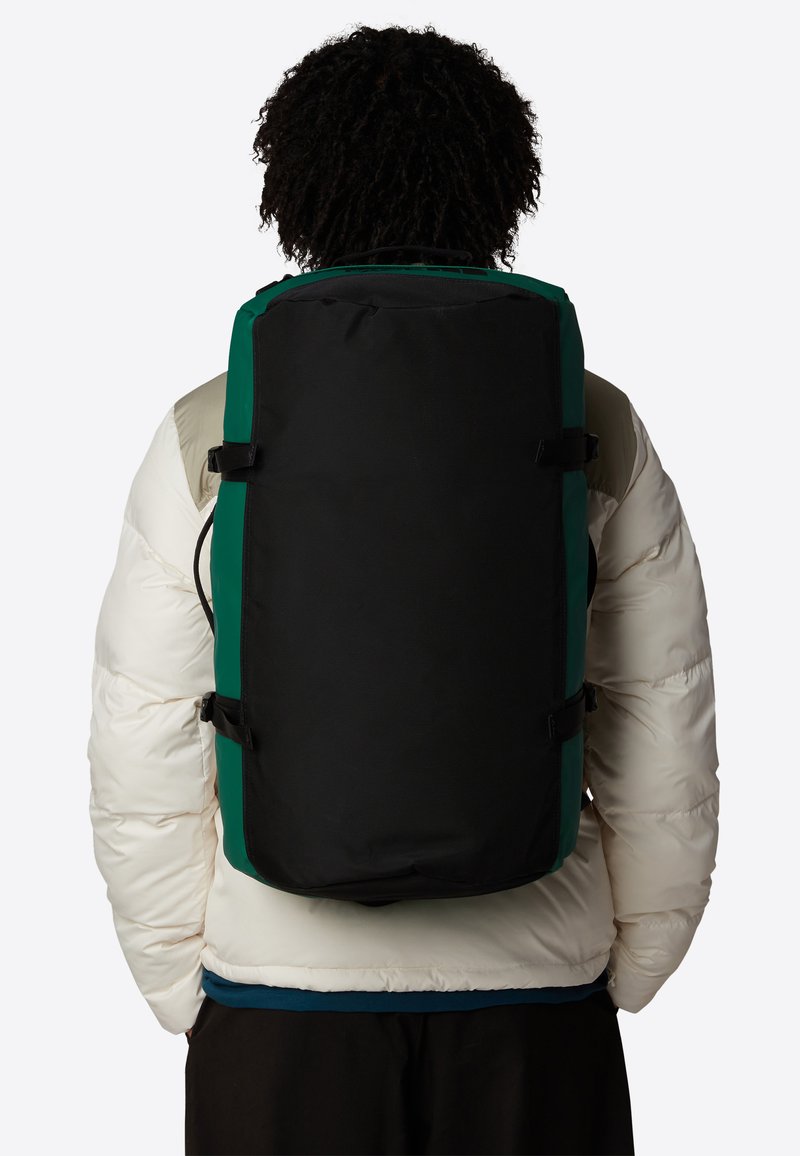 Sac à dos noir et vert de forme rectangulaire, en tissu, avec des bretelles ajustables et des boucles latérales, porté sur une veste blanche en duvet.