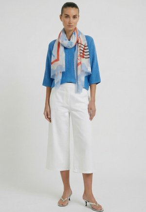 Vrouw draagt een blauwe gebreide top, witte broek met wijde pijpen en korte lengte, een meerkleurige sjaal en zilveren sandalen met hakken, staand tegen een effen achtergrond.