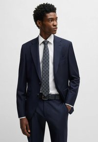 Abito blu navy con un design strutturato, camicia bianca e una cravatta scura a motivi. Presenta rever eleganti e una silhouette aderente.