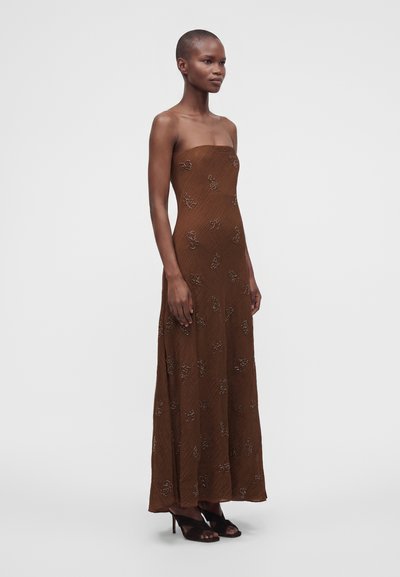 Femme portant une robe longue marron sans bretelles avec une broderie florale délicate et des sandales noires à talons hauts, debout devant un fond uni.