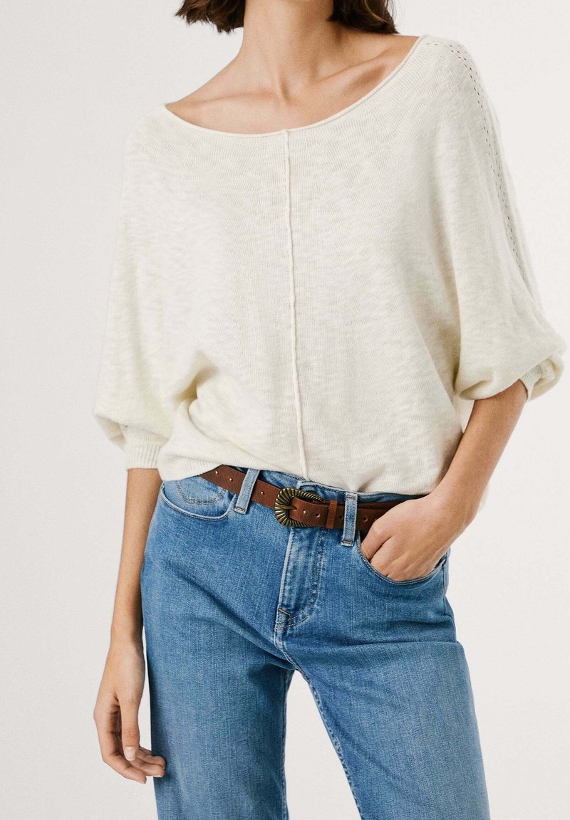 Femme portant un pull en maille blanc ample à manches mi-longues, rentré dans un jean bleu taille haute avec ceinture marron et boucle ornée.