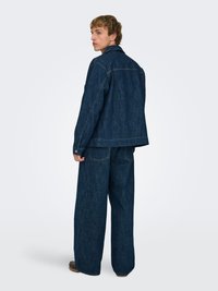 Denimjakke og vidbukser i mørkeblå. Kjennetegn inkluderer en klassisk krage, store lommer og kontrasterende sømmer.