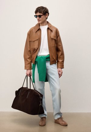 Fiatal férfi barna bőrdzsekiben, fehér ingben, világos farmerral és napszemüveggel, nagy sötétbarna táskát tart, amelyhez zöld pulóver van kötve deréknál.