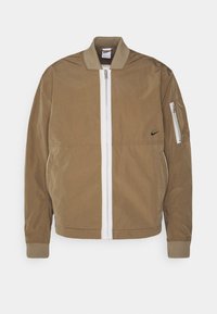 Brown bomber jacket with a white front zipper, ribbed cuffs, and a zippered sleeve pocket. Features a black Nike logo on the chest.