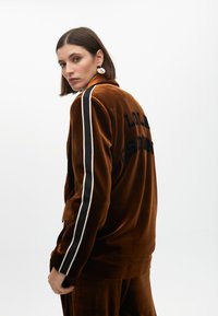 Brun sammetjacka med vitt och svart randdetaljer på ärmarna, med "LOLA" broderat på baksidan. Slät textur, avslappnad passform.