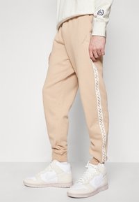 Beiga sweatpants med en vit sidrand som har ett broderat mönster, matchade med vita högklackade sneakers. Tillverkade av mjukt, strukturerat tyg.