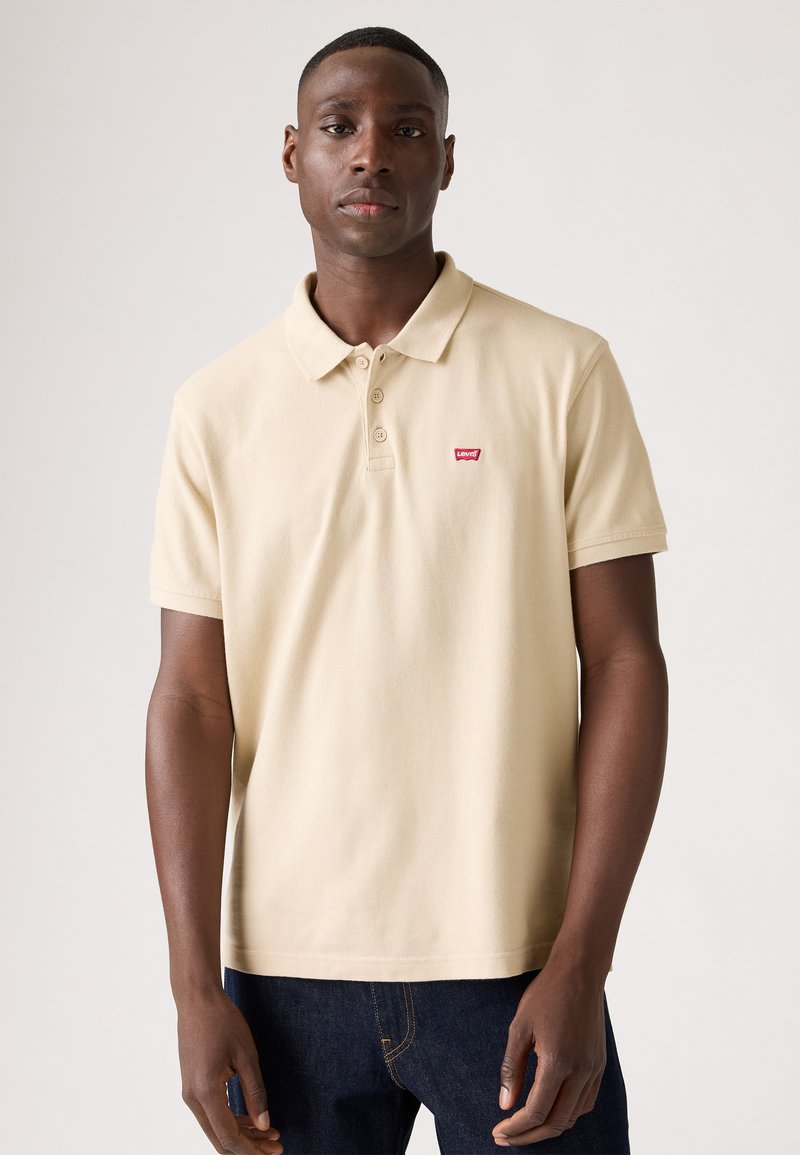 Camisa polo beige con cuello clásico, tres botones y un pequeño logo rojo de Levi's en el pecho; textura suave; combinada con vaqueros azul oscuro.