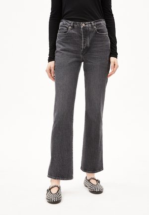 Person iført højtaljede sorte jeans og sorte Mary Jane-sko med nitter, stående mod en ensartet hvid baggrund.