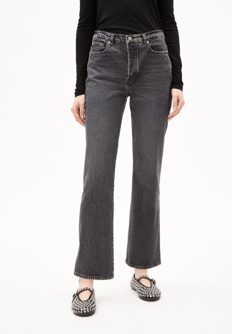 Personne portant un jean noir taille haute et des chaussures Mary Jane noires cloutées, debout devant un fond blanc uni.