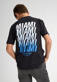 Camiseta negra con el texto repetido "MIAMI" en varias tonalidades de azul y blanco, con un logotipo en la parte inferior, llevada con pantalones grises.