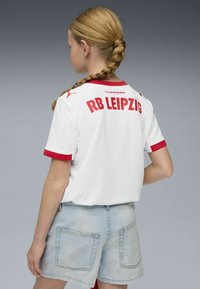Camiseta blanca con detalles rojos, con la inscripción "RB LEIPZIG" en letras negritas en la parte trasera, combinada con pantalones cortos de mezclilla azul claro. Textura suave.
