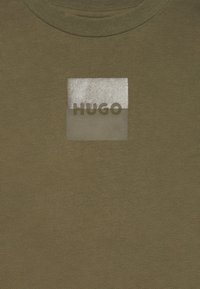 Olive zöld póló, melyen egy téglalap alakú ezüst és áttetsző folt található, rajta nagybetűs "HUGO" felirattal. Puha tapintású, kerek nyakkivágással.
