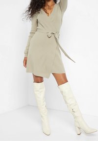 Nainen, joka on pukeutunut beigeen pitkähihaiseen kietaisumekkoon, jossa on vyötärölle sidottu nauha, yhdistettynä polveen asti ulottuviin valkoisiin korkokenkiin, seisoo valkoista taustaa vasten.