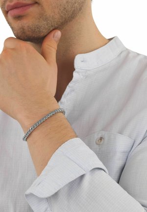 Uomo che indossa una camicia bianca con colletto alla coreana, toccando il mento con la mano sinistra, mostrando un braccialetto a catena d'argento al polso.