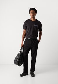 Men's black t-shirt with short sleeves, featuring a logo on the chest. Paired with tailored black pants and black leather shoes. Carrying a black duffel bag.