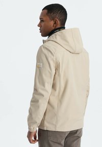 Beige mjukskaljacka med huva, har elastiska manchetter och ett sidoficka. Tyget har en jämn textur och är lämpligt för utomhusbruk.