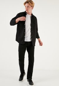 Crna košulja dugih rukava preko bijelog T-shirt-a, uparena s crnim hlačama i crnim tenisicama. Košulja ima džep na prsima i opušteni kroj.
