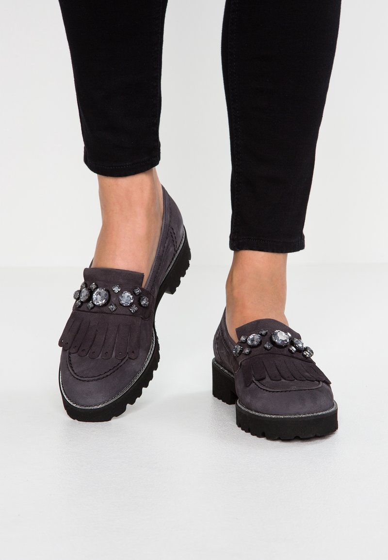Mocassins en daim gris foncé avec semelles noires épaisses et grandes pierres précieuses transparentes en embellissement sur le dessus, portés avec un pantalon noir ajusté.