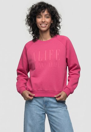 Frau mit lockigem Haar lächelt, trägt ein knallrosa Sweatshirt mit dem Schriftzug "ALIFE" und hellblaue Jeans, steht vor einem einfarbigen Hintergrund.