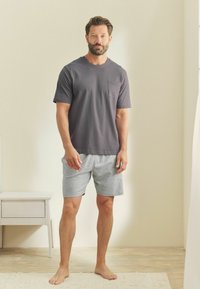 Kortärmad grå t-shirt för män med ficka, i kombination med ljusgrå shorts, båda i mjukt, andningsbart material. Modell som står på ett bar golv.